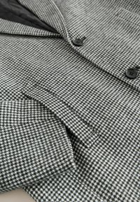 Sivý a čierny kabát s vzorom houndstooth má vystúpený golier, vrecko s chlopňou a čierne gombíky na textúrovanej látke.