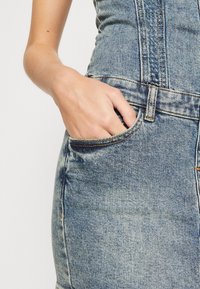 Džínové šaty se světlým modrým odstínem, strukturovaným střihem, viditelným prošitím a přední kapsou, do které je vložena ruka pro detail.