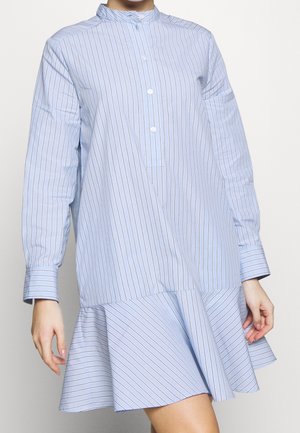 Vrouw draagt een lichtblauwe jurk met dunne verticale strepen en een asymmetrische uitlopende zoom, een voorzijde met knopen en lange mouwen met manchetten.