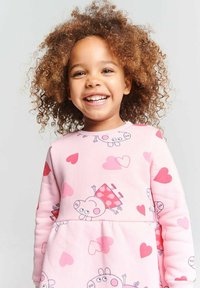 Lächelndes junges Mädchen mit lockigem Haar, das ein rosa Kleid trägt, das mit Cartoon-Schweinen und roten sowie rosa Herzen verziert ist, vor einem einfarbigen Hintergrund.