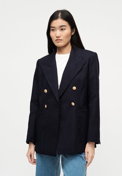 Νεαρή γυναίκα με μακριά σκούρα μαλλιά, ντυμένη με μπλε διπλό παλτό με χρυσά κουμπιά και ανοιχτό γαλάζιο τζιν, στέκεται μπροστά σε λευκό φόντο.