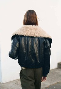 Giacca nera in pelle cropped con un soffice colletto in shearling beige, caratterizzata da spalle imbottite e texture liscia. Indossata con pantaloni verde oliva.