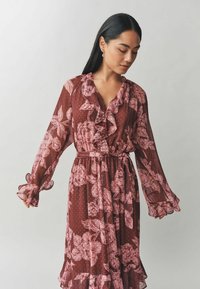 Ilga burgundi spalvos suknelė su rožinėmis gėlių raštais, volanais ir permatomomis rankovėmis. Prisegama diržu liemens vietoje, su taškinėmis tekstūromis.