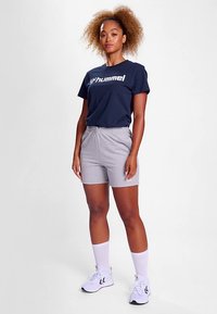 Marinblå t-shirt med vit logotyp, kombinerad med ljusgrå shorts. Modellen bär vita strumpor och sneakers. Avslappnad sportklädsel.