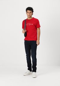 Raudona medvilninė marškinė su "TOMMY HILFIGER EST. 1985" grafika, derinami su tamsiai mėlynomis džinsinėmis kelnėmis ir baltomis sportbačiais. Paprastas dizainas, kasdienis siluetas.