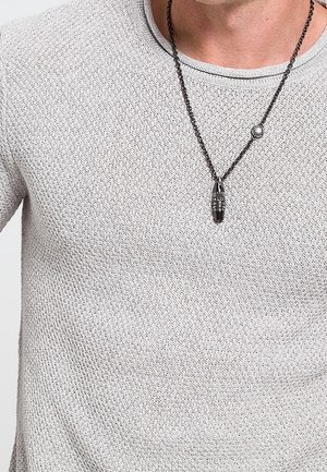 Homme portant un pull en maille texturée gris clair et un collier chaîne noir avec un pendentif cylindrique noir.