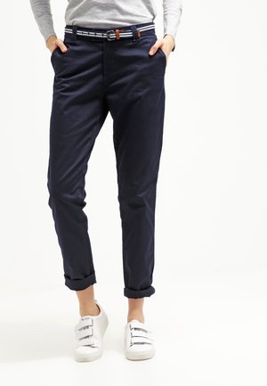 Personne portant un pantalon bleu marine retroussé avec une ceinture rayée et des baskets blanches, les mains dans les poches, debout devant un fond blanc.