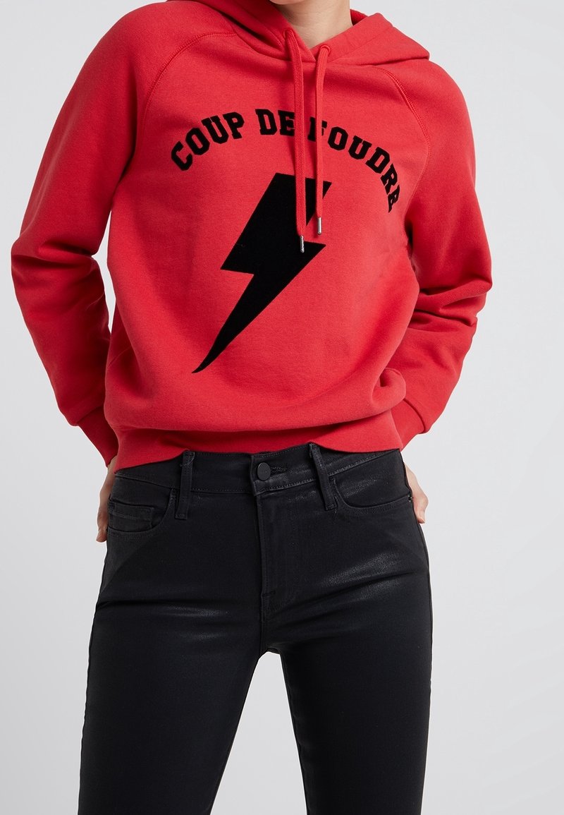Personne portant un sweat à capuche rouge avec un éclair noir et le texte "COUP DE FOUDRA", associé à un pantalon noir taille haute.