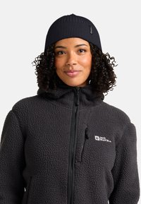 Donna con capelli ricci che indossa un berretto a coste nero e una giacca in pile grigia scura con zip, con il logo Jack Wolfskin sul petto.