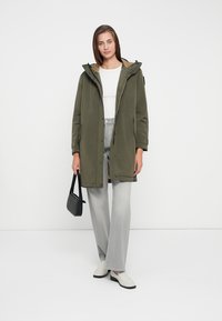 Olive zaļš parka ar kapuci, ar izmantojamu rāvējslēdzēju priekšpusē un sānu kabatām. Saskaņots ar pelēkām platām biksēm un baltām slip-on kurpēm.