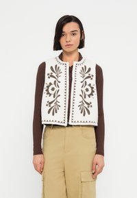 Gilet en polaire ivoire avec broderie florale brun foncé et bords festonnés. Doté d'un col rond et d'une ouverture frontale. Porté par-dessus un haut marron.