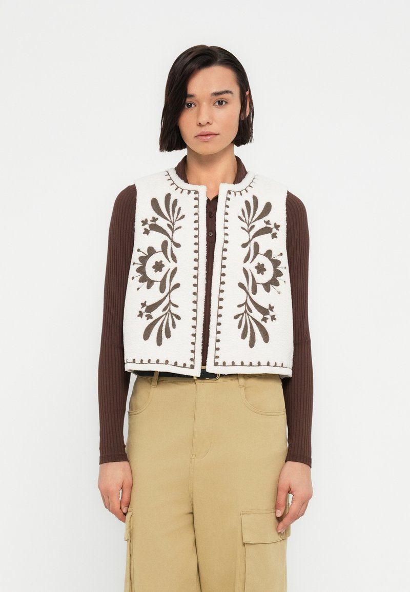 Gilet en polaire ivoire avec broderie florale brun foncé et bords festonnés. Doté d'un col rond et d'une ouverture frontale. Porté par-dessus un haut marron.