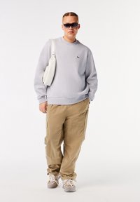 Felpa grigia con logo, pantaloni cargo beige e sneakers grigie chiare. Accessorio: borsa a tracolla bianca. Sfondo bianco uniforme.