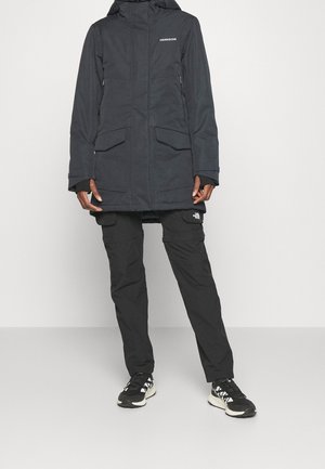 Personne portant un parka à capuche noir Didriksons, un pantalon cargo noir de The North Face et des baskets noires et blanches, debout devant un fond uni.
