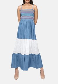 Vestido maxi de algodón azul con cuerpo smock, detalles de ojales blancos y tirantes finos. Se complementa con tacones nude.