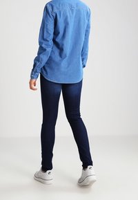 Camisa de denim azul claro con puños abotonados, jeans ajustados azul oscuro y zapatillas blancas, exhibidos desde la espalda contra un fondo liso.