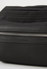 Calvin Klein juodas audinio maišelis su tekstūruotu paviršiumi, išskirtiniu sidabriniu užtrauktuku ir subtiliu prekės ženklu šone.