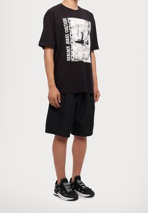 Person trägt ein schwarzes, übergroßes Versace Jeans Couture T-Shirt mit Grafikdruck und schwarze, lockere Shorts, kombiniert mit schwarz-weißen Schnürschuhen.
