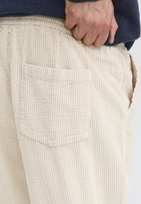 Beżowe spodnie sztruksowe o fakturze żebrowanej, z boczną kieszenią i marszczonym paskiem w talii. Zbliżenie podkreśla miękkość materiału.
