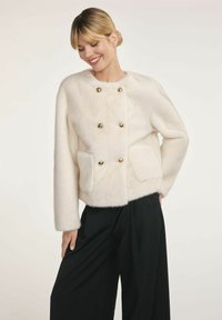 Uśmiechająca się kobieta w białym, puszystym, podwójnym płaszczu zapinanym na złote guziki i czarnych szerokich spodniach na jednolitym tle.