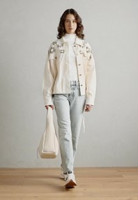 Krembrodert jakke over en hvit turtleneck, lyseblå jeans og hvite sneakers, bærende en beige håndveske. Bakgrunn med tregulv.