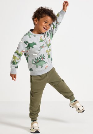 Grijze sweatshirt met dinosaurustafereel in groen, geel en rood; gecombineerd met olijfgroene joggingsbroek en beige sneakers.