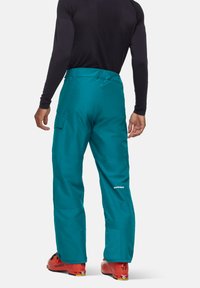 Mann trägt tealfarbene Cargo-Hosen, ein schwarzes langärmliges Shirt und rote Skischuhe, steht vor einem schlichten Hintergrund und blickt weg.