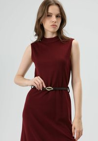 Bordeauxrode mouwloze jurk met een geribbelde textuur, voorzien van een hoge halslijn en een zwarte riem met een gouden accent in de taille.