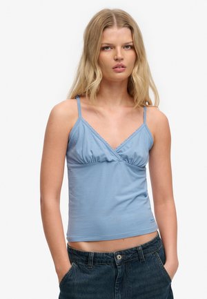 Młoda kobieta o długich blond włosach, ubrana w jasnoniebieski top na cienkich ramiączkach i ciemnoniebieskie dżinsy, z rękami w kieszeniach.