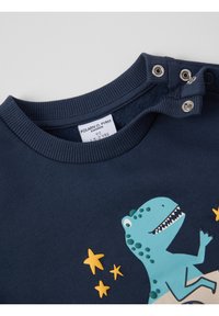 Navyblauwe kindershirt met drukknoopjes op de schouder, met een glimlachende blauwe dinosaurus en drie gele sterillustraties.