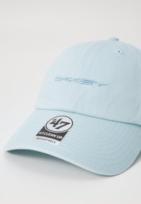 Căciulă de culoare albastru deschis, confecționată din material textil, cu un logo brodat și o curea ajustabilă. Include un eticheta alb-negru cu instrucțiuni de îngrijire.