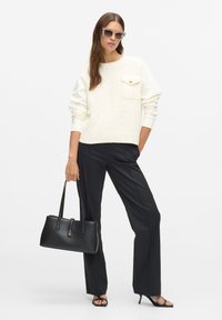 Krémový sveter s textúrou, vreckom a zlatým gombíkom, v kombinácii s čiernymi nohavicami na mieru a čiernou strukturovanou kabelkou. Priehľadné slnečné okuliare.