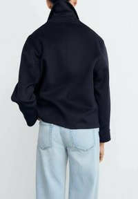Persona che indossa una giacca nera a colletto alto e jeans azzurri chiari, vista da dietro su uno sfondo chiaro e semplice.