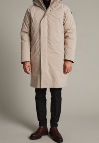 Beige vattentät parka med hög krage, dragkedjestängning, framfickor och huva. Bärs över svarta byxor och bruna texturerade kängor.