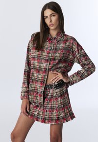 Karierter Hemd in Rot, Schwarz und Beige mit Knopfleiste und Brusttaschen, kombiniert mit einem gefalteten Rock und einem schwarzen Gürtel mit silbernen Ösen.