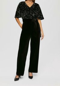 Svart sammetsoverall med en paljettklädd topp med kort ärm, V-ringning och knytmidja. Kompletteras med svarta högklackade skor.