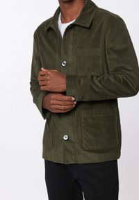 Zielonooliwkowa kurtka z segmentu corduroy z kołnierzem, posiadająca kieszeń na piersi i dwie kieszenie z przodu, zapinana na czarne guziki. Proste mankiety rękawów.