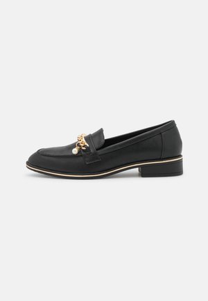 Zwarte leren loafer met lage hak, gouden ketting en pareldetail bovenop, en een dunne gouden rand boven de zool.