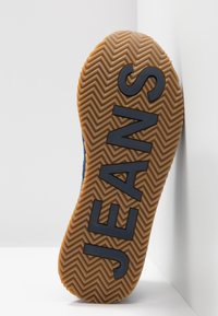 Semelle de sneaker en caoutchouc brun avec un motif en chevrons. Présente des lettres noires en gras écrivant "JEANS." Le côté montre un dessus en textile bleu.