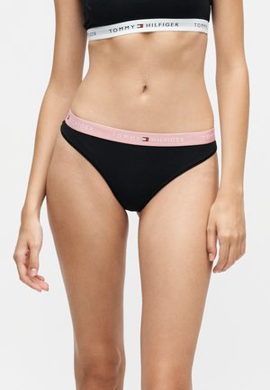 Kobieta nosząca czarne bieliznę Tommy Hilfiger z różową gumką i czarny sportowy biustonosz z białą gumką, oba z logo Tommy Hilfiger.