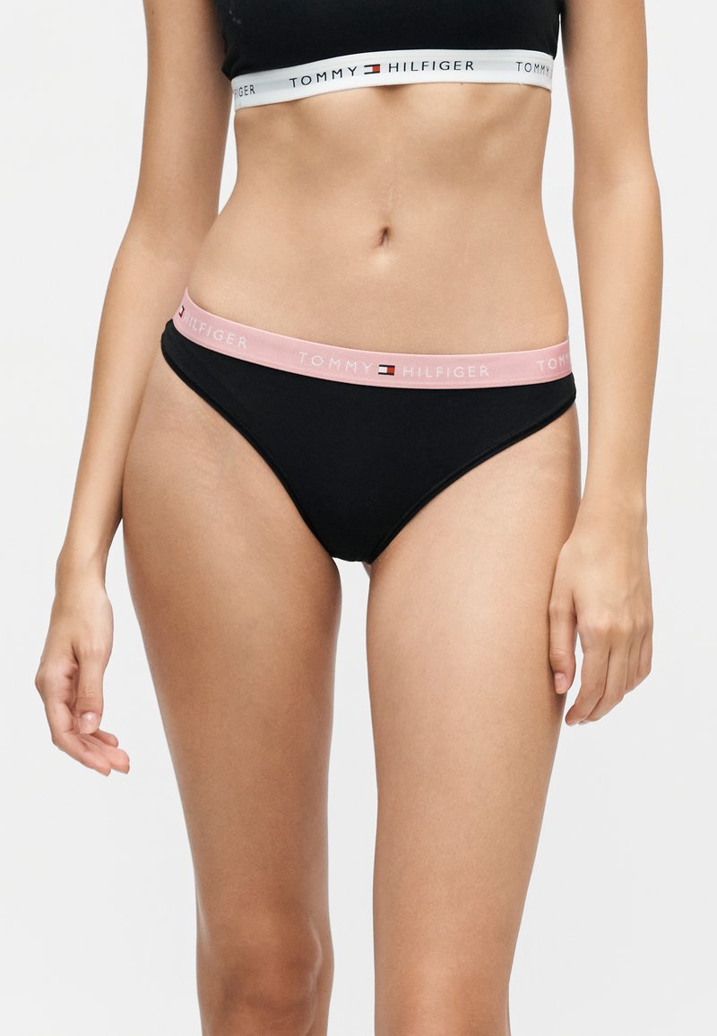 Femme portant des sous-vêtements noirs Tommy Hilfiger avec une ceinture rose et un soutien-gorge de sport noir avec une ceinture blanche, tous deux portant le logo Tommy Hilfiger.