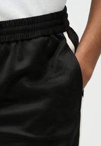 Μαύρο αθλητικό σορτς με ελαστική μέση και λευκές ρίγες στα πλευρά, χέρι στην δεξιά τσέπη, διακριτικό λογότυπο "G P H STUDIOS" μπροστά.