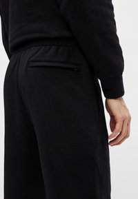 Juodos sportinės kelnės, pagamintos iš medvilnės mišinio; turi elastinę juosmenį ir užtrauktuku užsegamą kišenę gale, su lygiu paviršiumi.