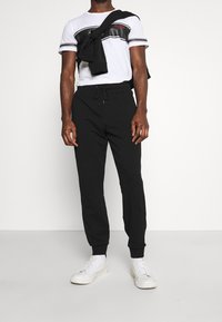 Mann in schwarzen Jogginghosen, weißen Sneakers, einem weiß-schwarz gestreiften T-Shirt und einem schwarzen Sweatshirt, das um seine Schultern gebunden ist.