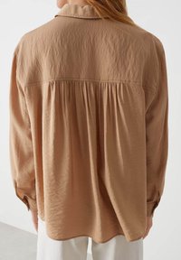 Camicia a maniche lunghe color beige chiaro con colletto a risvolto e dettagli arricciati sulla schiena. Tessuto morbido e testurizzato con vestibilità ampia e polsini con bottoni.