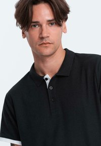 Zwarte poloshirt met een witte kraag, voorzien van drie knopen en een gestructureerde stof. Het ontwerp heeft korte mouwen en een sportieve uitstraling.