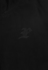 Czarny t-shirt z materiału, z subtelnym, wytłoczonym logo na środku. Materiał wydaje się miękki, o gładkiej fakturze i bez widocznych wzorów.