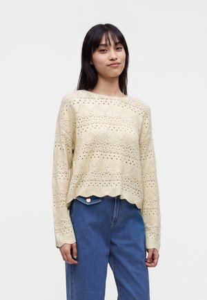 Ung kvinde med glat sort hår iført en cremefarvet strikket sweater med tekstureret mønster og blå højtaljede jeans mod en ensfarvet baggrund.