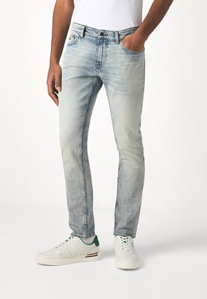 Uomo che indossa jeans aderenti azzurri chiari e sneakers bianche con dettagli verdi, in piedi davanti a uno sfondo bianco semplice.