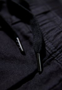 Zbliżenie czarnych spodni z materiału, z czarnymi wplecionymi końcówkami sznurków i szczegółowym przeszyciem na pasie.